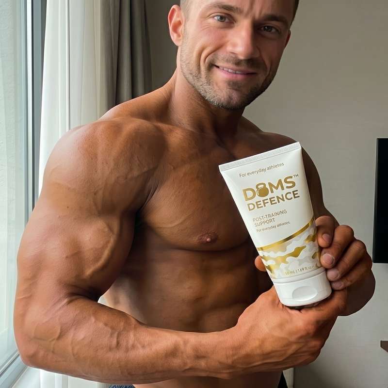 Muscular man holding a 'Doms Defence' muscle cream product in a room with a window.